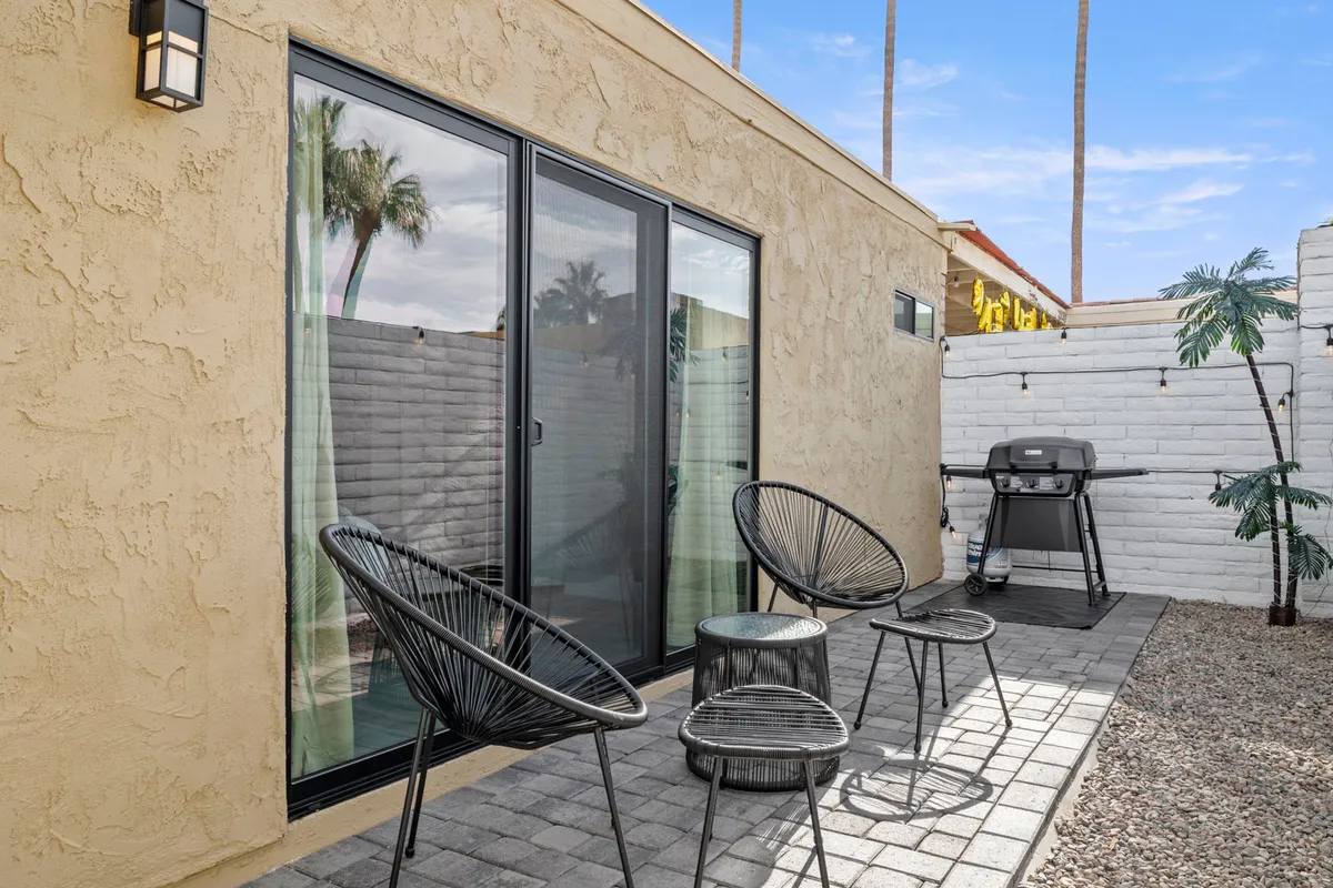 A back patio scene featuring glass doors, comfortable black chairs, a grill, and palm trees, evoking a sense of privacy and comfort ideal for alfresco dining or watching outdoor TV. The layout invites gatherings or solo relaxation in a sheltered backyard setting.