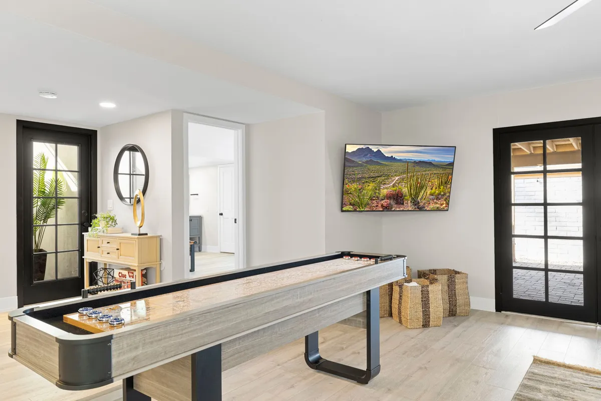 A spacious interior game room featuring a shuffleboard table, a large TV displaying a landscape image, black-framed glass doors opening to the outside, and a decorative basket. The room is bathed in natural light, blending indoor comfort with outdoor views.