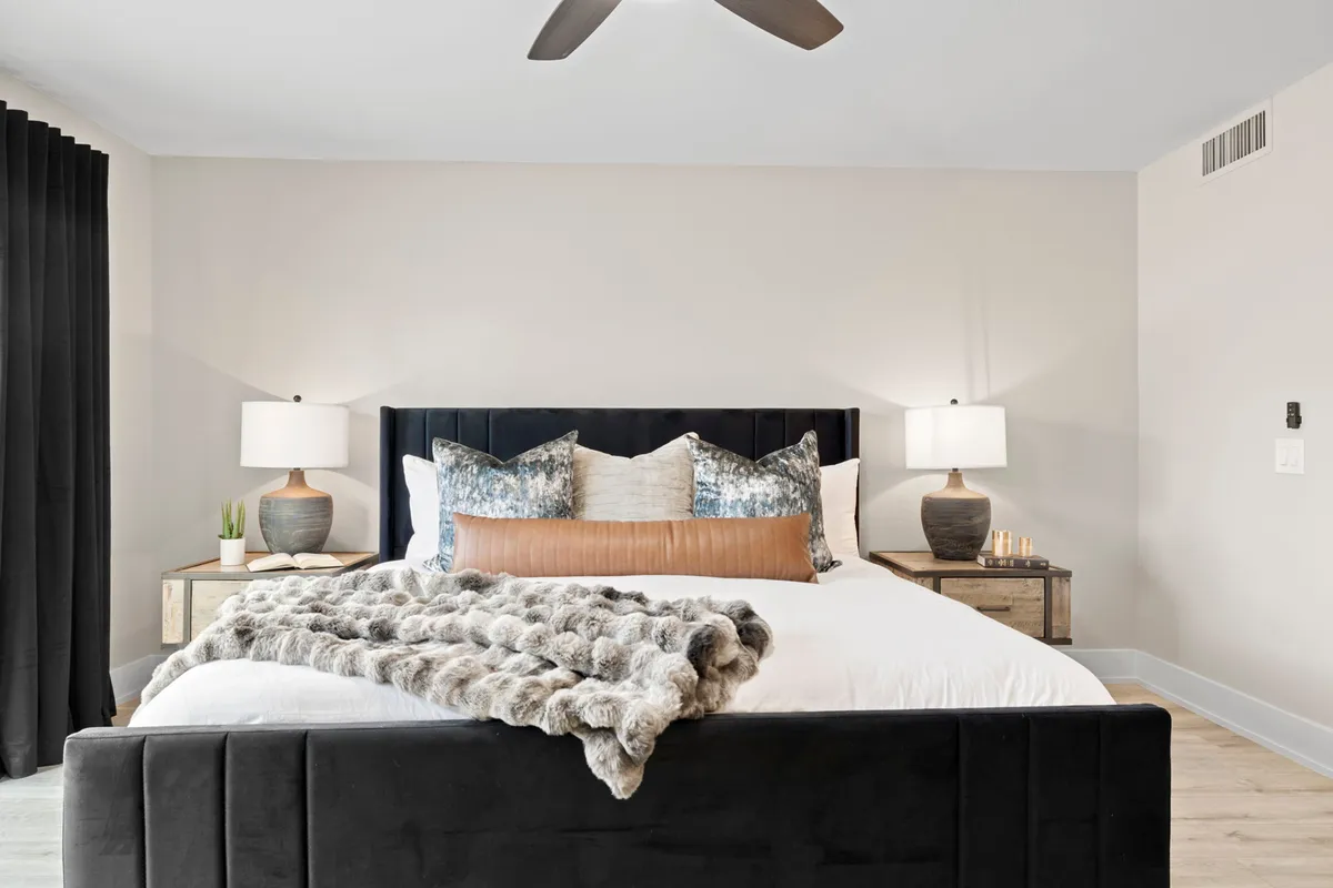 This image features a bed with plush pillows and a soft throw blanket, flanked by nightstands with lamps, and curtains framing a sunlit window. The mood is relaxing and cozy, inviting guests to unwind and enjoy restful sleep.