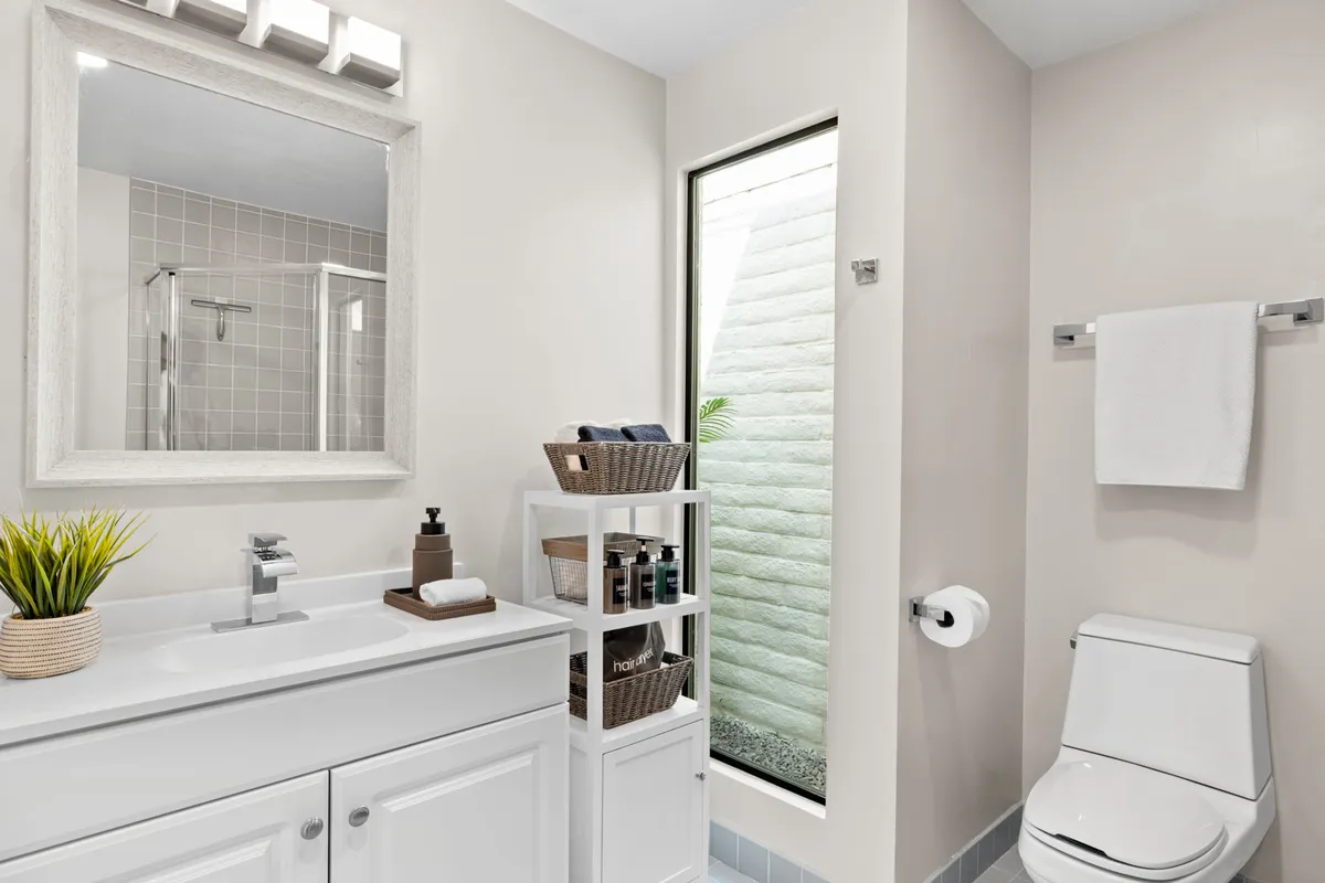 A bright, inviting bathroom scene featuring a sleek vanity with sink, a large mirror, and storage shelving. A window offers greenery views, and the organization suggests high-end toiletries and ample storage for extended stays.