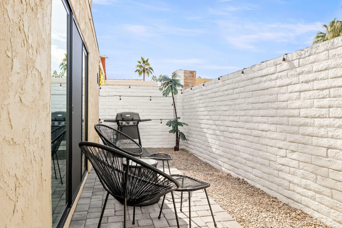 An inviting outdoor patio setup featuring black chairs, a barbecue grill, palm trees, and a white privacy wall. The atmosphere feels relaxed and social, with natural greenery and plenty of space for gatherings or a quiet morning coffee.