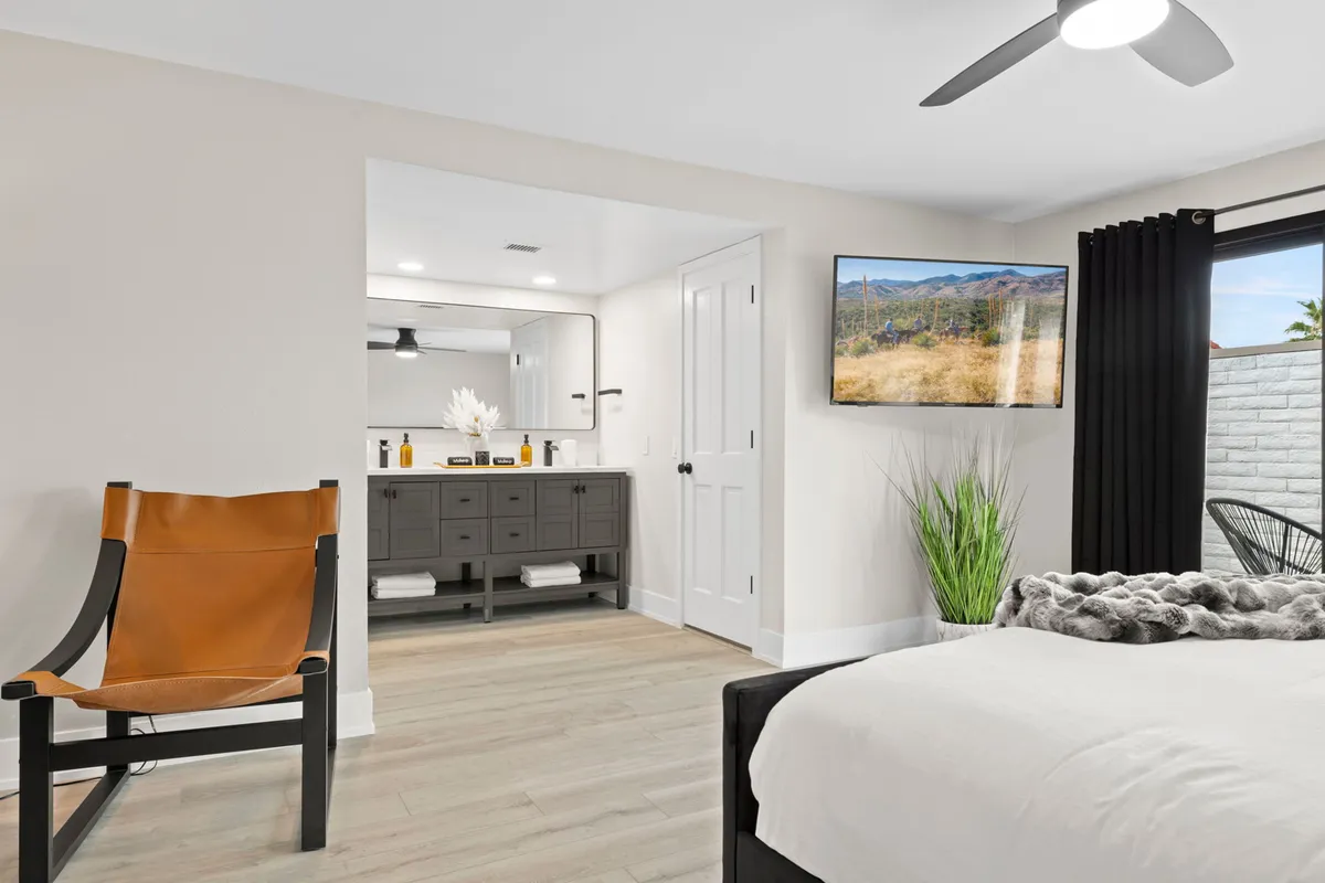 A serene bedroom scene with a cozy bed, a comfortable chair, vanity, television, and a bright window view. The ambiance is peaceful and inviting, ideal for unwinding after a productive day or taking a mid-afternoon break.