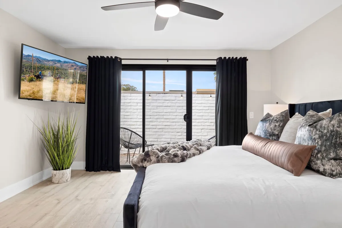 A tranquil king-sized bed with decorative pillows sits beneath a large television mounted on the wall. Indoor plants and black curtains enhance the cozy, modern atmosphere, while natural light softly fills the space.