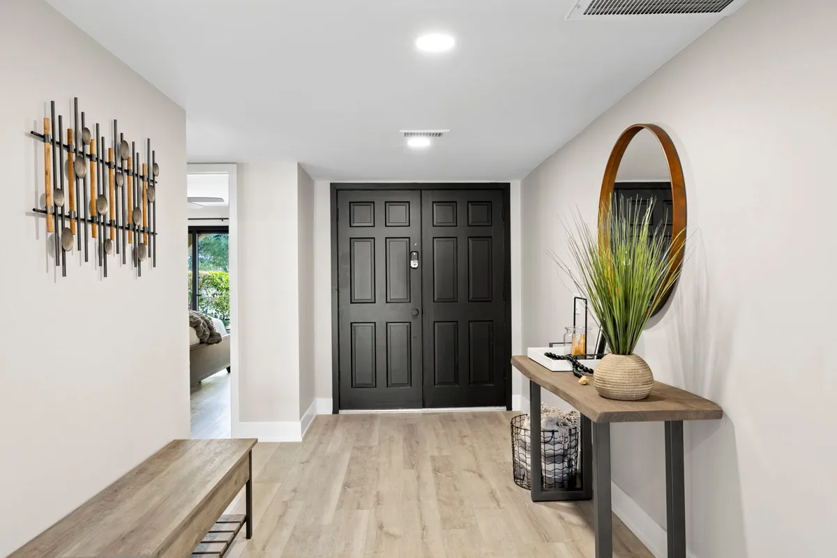 A modern and inviting foyer area featuring a black front door, decorative wall art, a console table with thoughtfully placed décor, and a potted plant. The space is well-lit, offering a sense of warmth and style immediately upon entering.