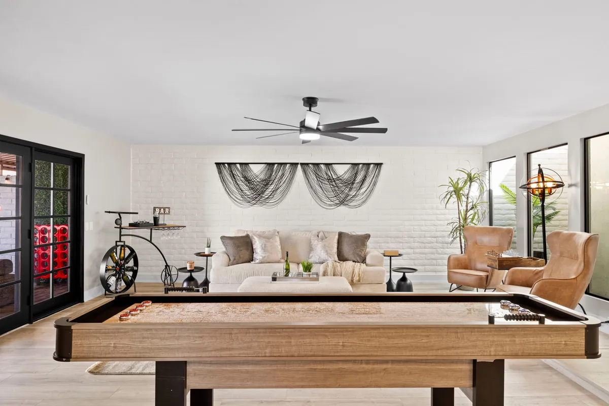 A vibrant, sunlit room featuring a shuffleboard table, stylish armchairs, a comfortable sofa, large windows that let in natural light, and decorative wall art. The space exudes a playful, family-friendly atmosphere ideal for indoor games and social gatherings.